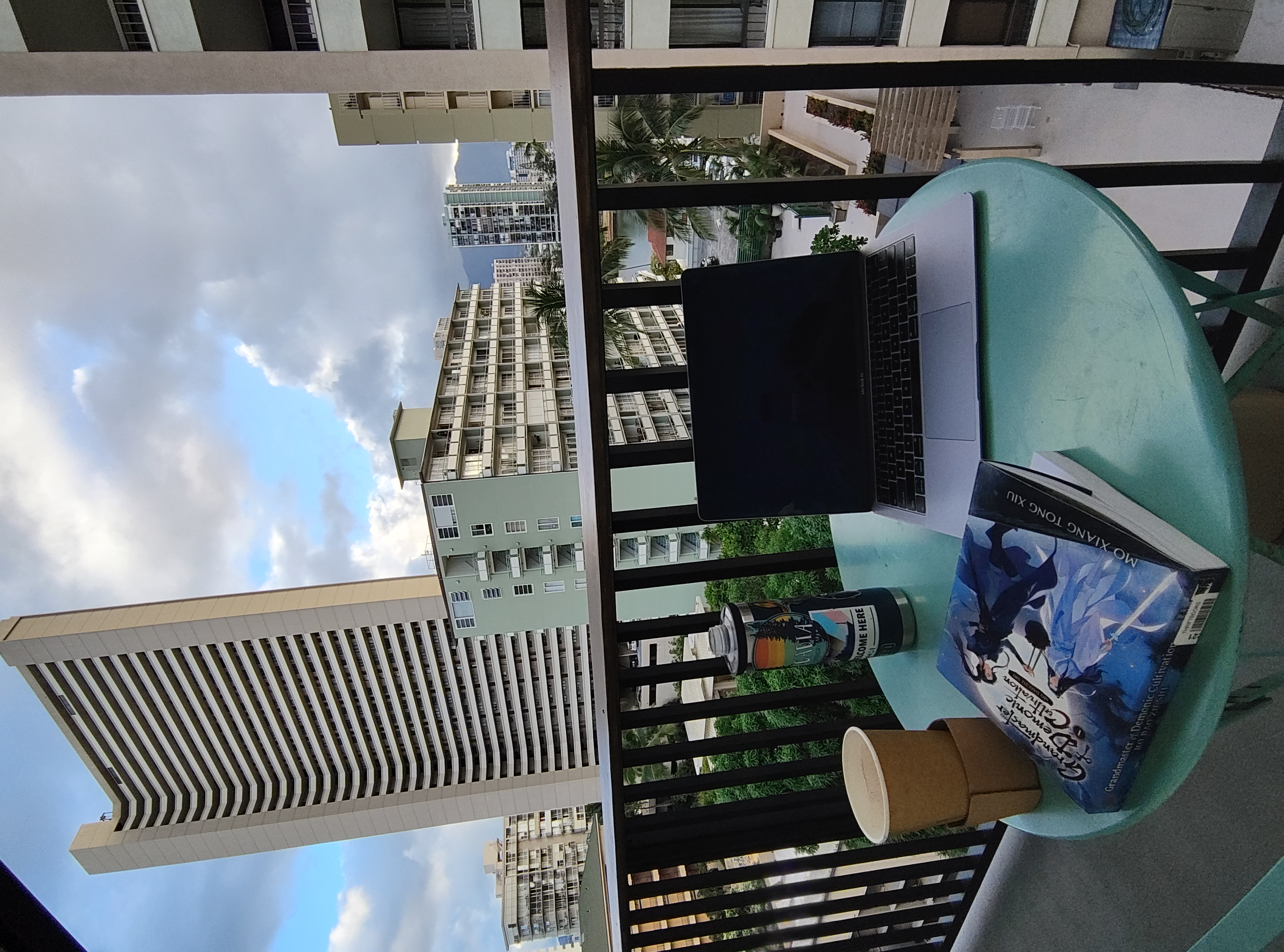 a picture taken on a hotel balcony. There is a small table with an empty take out coffee cup, a laptop, a water bottle, and a book. In the background are a blue sky with fluffy white clouds and tall, light colored hotel and apartment buildings.